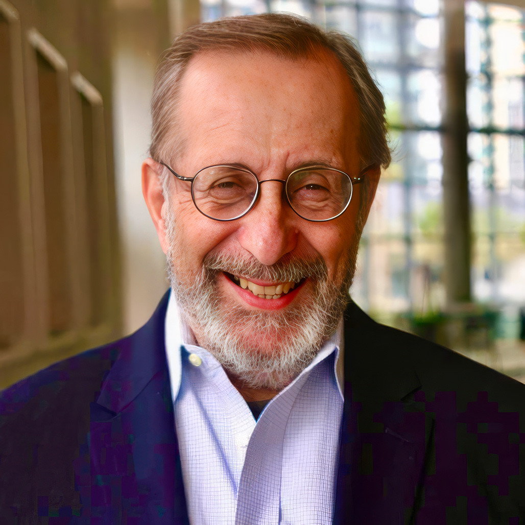 A portrait of smiling older man John DeDakis with a gray beard and round glasses, wearing a dark blue blazer over a light blue collared shirt. The softly blurred modern indoor background features large windows. This image relates to the topic  Can Writing Really Help with My Grief?    GPS Hope - Grieving Parents Sharing HOPE.