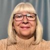 Close-up of a smiling grieving parent with blonde hair, bangs, and tortoiseshell glasses, wearing a beige turtleneck sweater, representing warmth and compassion. Honoring Our Children Who Died by Suicide. GPS Hope – Grieving Parents Sharing HOPE.